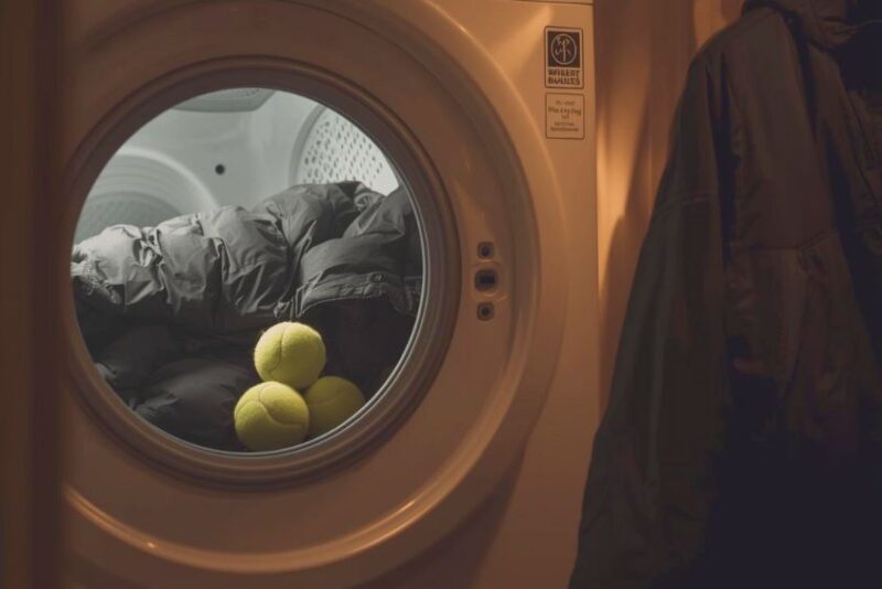 A down jacket inside a tumble dryer with two clean tennis balls. Next to it, a synthetic jacket hanging on a drying rack.